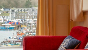 A red sofa with a patterned pillow, a window with a view of a harbor, and a building with the sign "FATTACE". - Jetty Cottage by Interhome (Mevagissey)