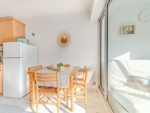 A compact kitchen with a white refrigerator, wooden cabinets, and a dining table with chairs. A decorative sunburst wall art hangs above the table. - du Parc II by Interhome (La Grande Motte)