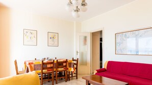 Una sala de estar con una mesa de comedor de madera, sillas, un sofá rojo y un sillón amarillo. Hay dos cuadros en las paredes y un mapa colgado en la pared.