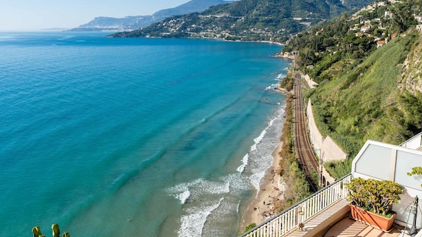 Vista costera con una vía de tren que corre paralela a la playa, un cactus en primer plano y un balcón con plantas en macetas.