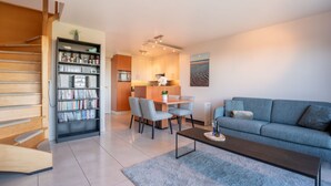 A modern living room with a sofa, coffee table, and a bookshelf. There is a dining area with a table and chairs. The room has a staircase and a kitchen in the background. - Marebelle with seaview by Interhome (Bredene)