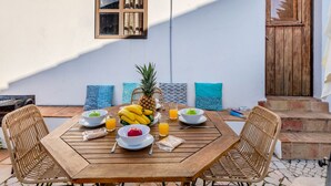 Une table en bois, dressée avec des fruits et des boissons, des chaises en osier et un mur blanc avec une fenêtre et une porte.
