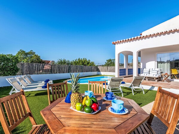 Une terrasse avec une table en bois dressée pour un repas, une piscine et des sièges d’extérieur.