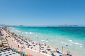 Een strand met helder turquoise water, parasols en mensen.