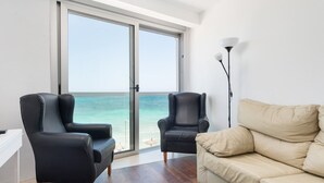 Un salon moderne doté d’une grande porte-fenêtre coulissante qui donne sur un balcon offrant une vue sur l’océan. On y trouve deux fauteuils en cuir noir et un canapé beige.