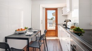 Une cuisine moderne avec des carreaux blancs, une porte en bois donnant sur un balcon, une table à manger avec des chaises et un plan de travail sur lequel se trouvent un grille-pain et des légumes frais.