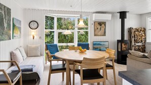 A cozy living room with a round wooden table, blue cushioned seating, a fireplace, and large windows offering a view of greenery.