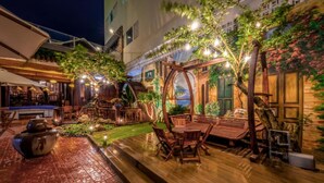 Un patio al aire libre con mobiliario de madera, luces colgantes y vegetación.
