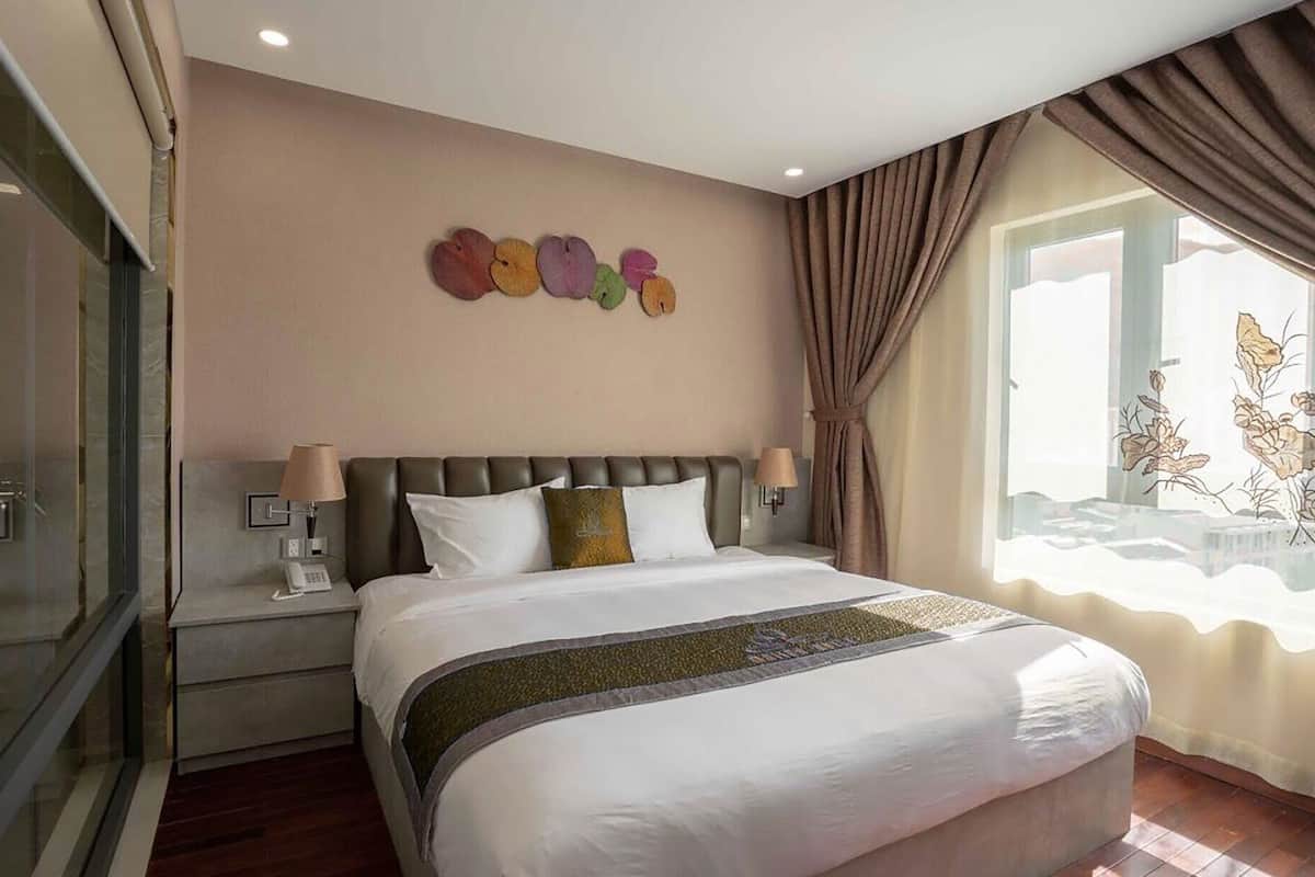 Habitación de hotel con una cama grande, una mesita de noche con lámpara, un reloj en la pared y una ventana con cortinas.