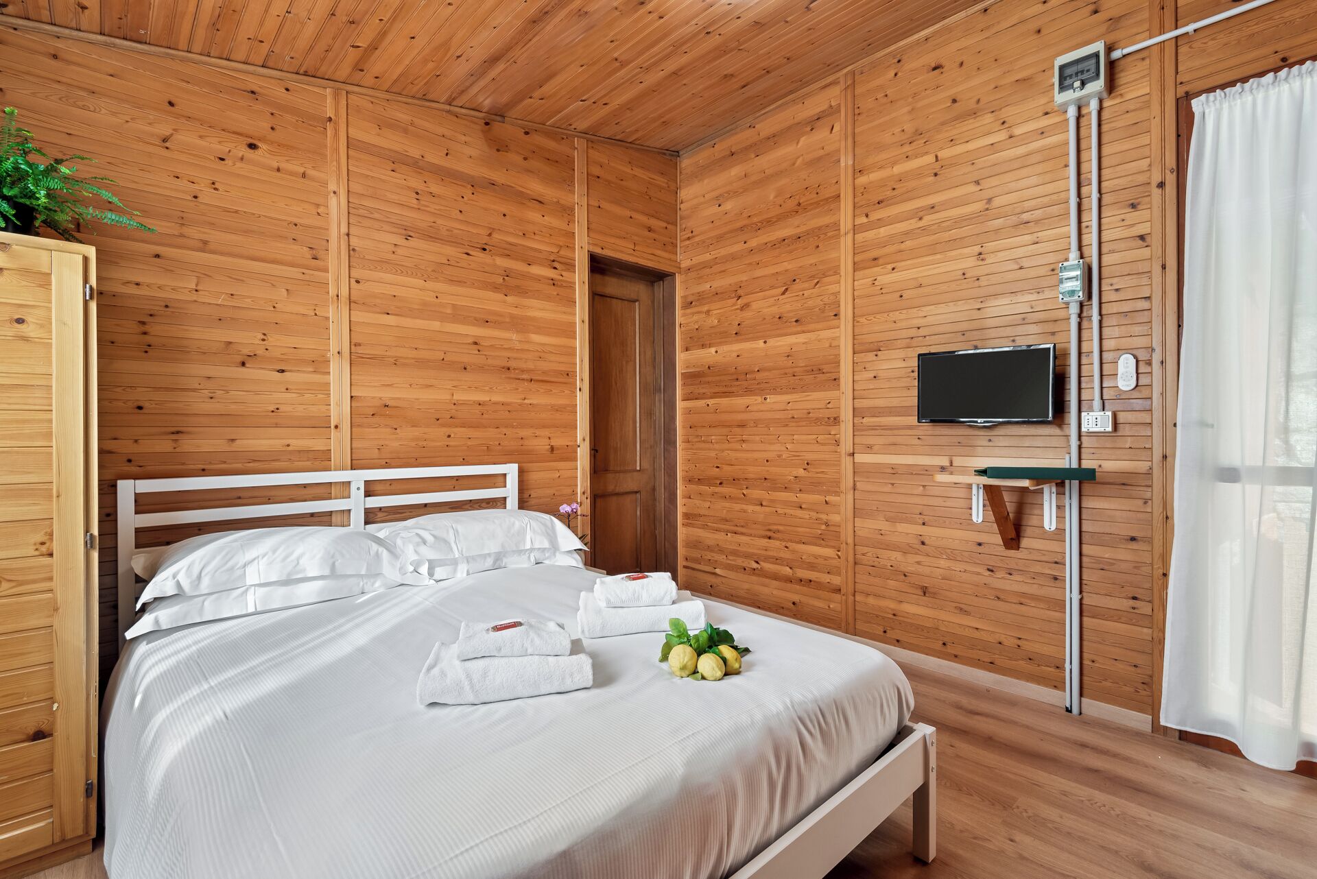 Um quarto com painéis de madeira, uma cama, televisão na parede e uma janela com cortinas.