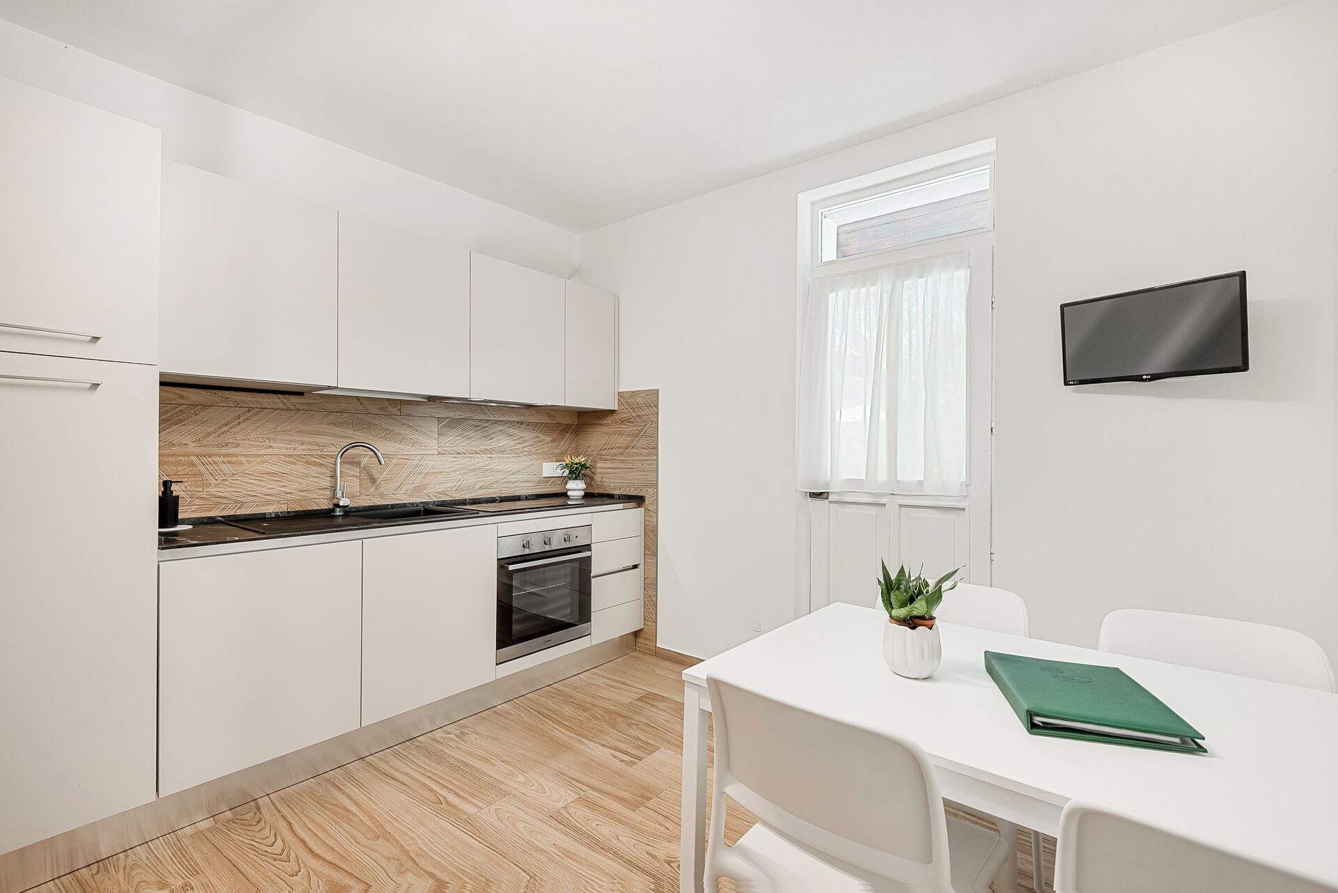 A modern kitchen with white cabinets, a black countertop, and a built-in oven. There is a dining table with chairs and a green book on it. A TV is mounted on the wall.