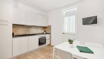 A modern kitchen with white cabinets, a black countertop, and a built-in oven. There is a dining table with chairs and a green book on it. A TV is mounted on the wall.