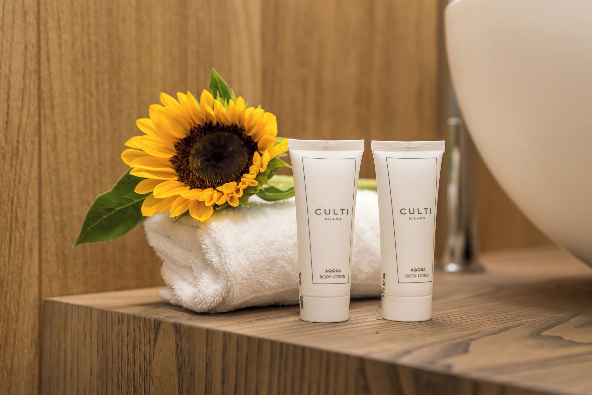 A bathroom with a wooden vanity, two tubes of Culti Milano body lotion, a white towel, and a sunflower.