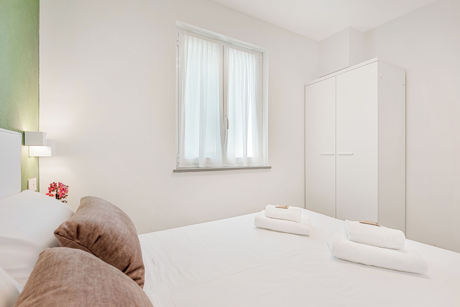 A hotel room with a bed, a wardrobe, a window with curtains, and a bedside table with a lamp and flowers.