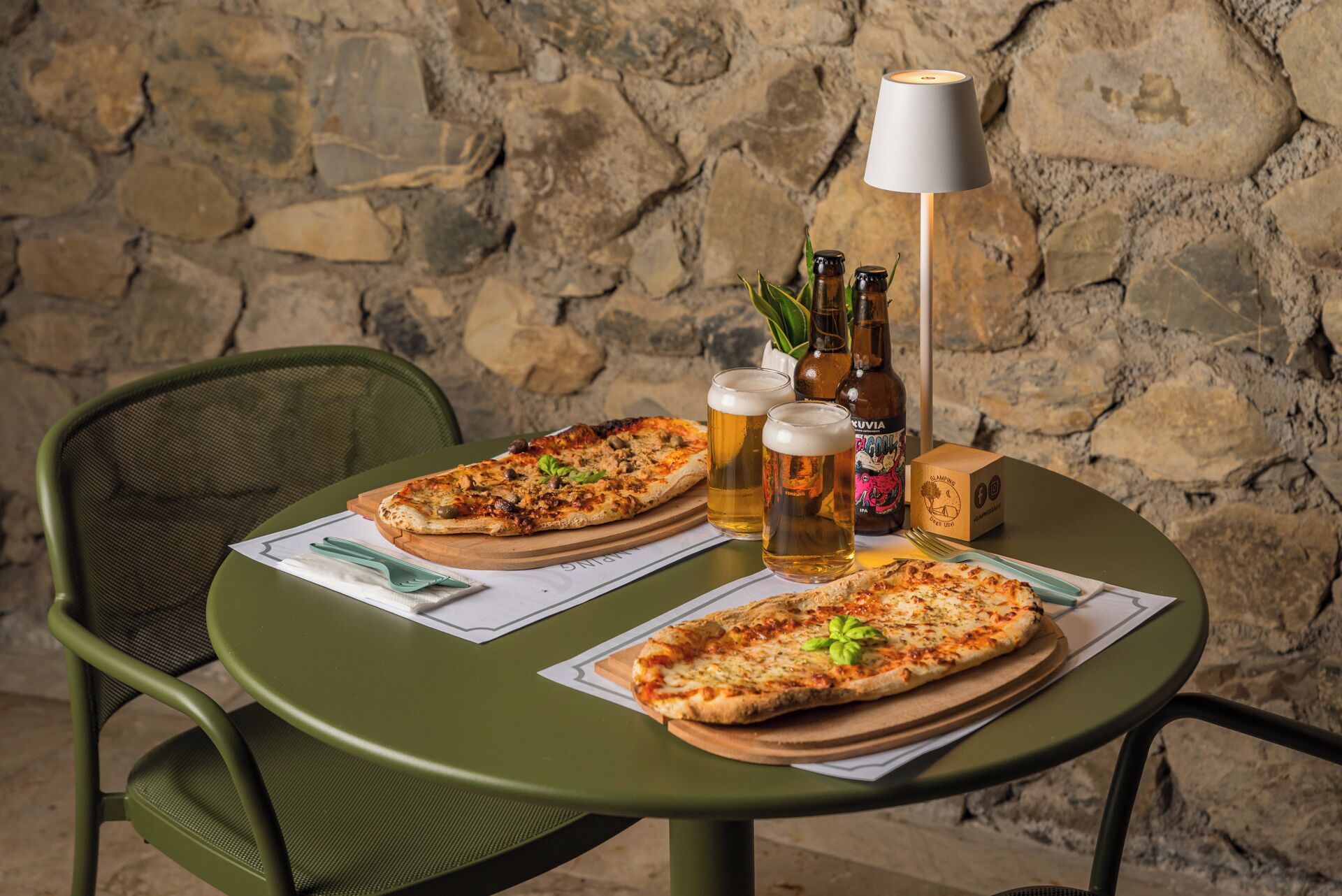Två pizzor på ett runt bord, tillsammans med en lampa, två ölglas och en ölflaska.