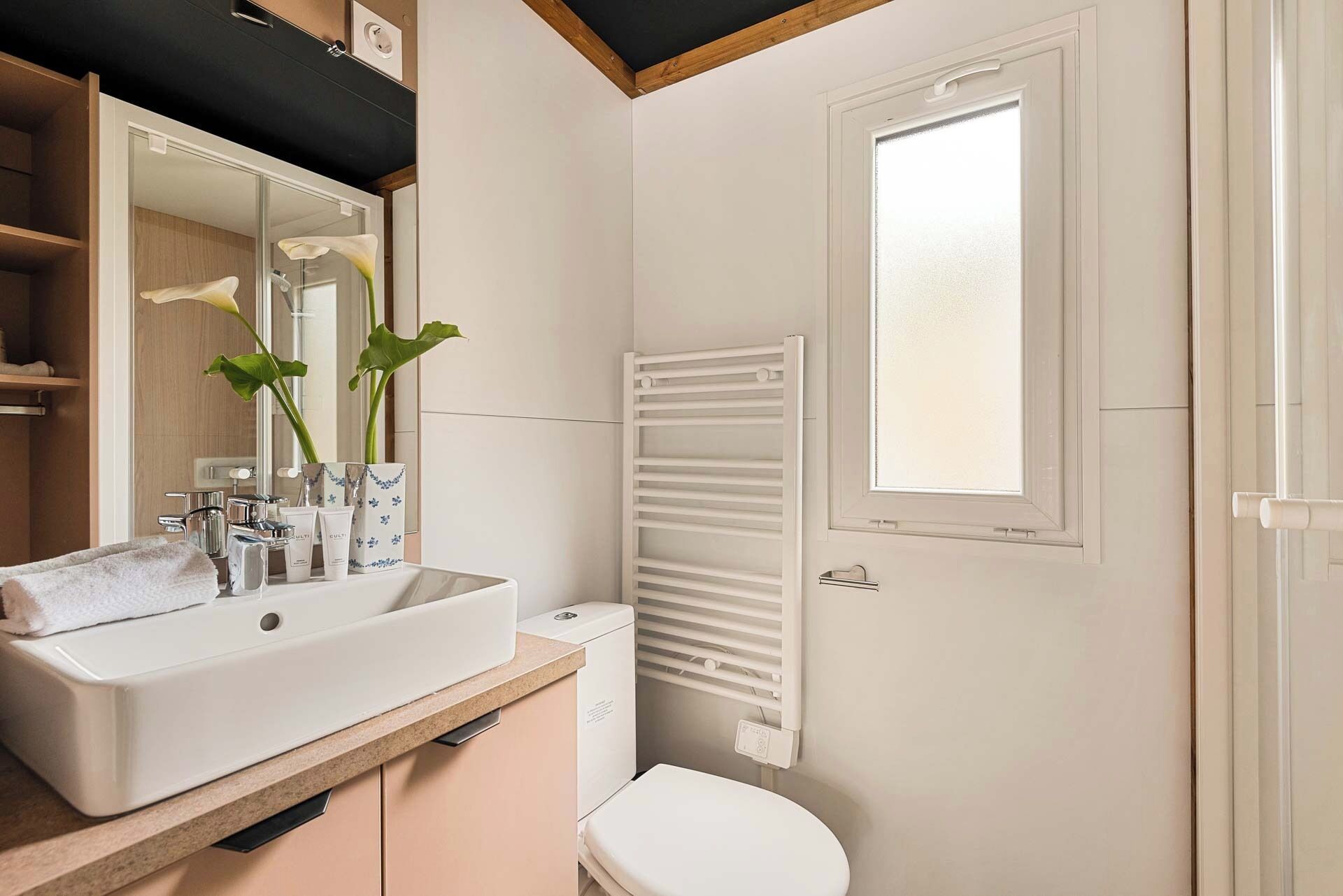 Une salle de bain avec un lavabo blanc, un miroir et des toilettes. Il y a une fenêtre avec des stores et un porte-serviettes.