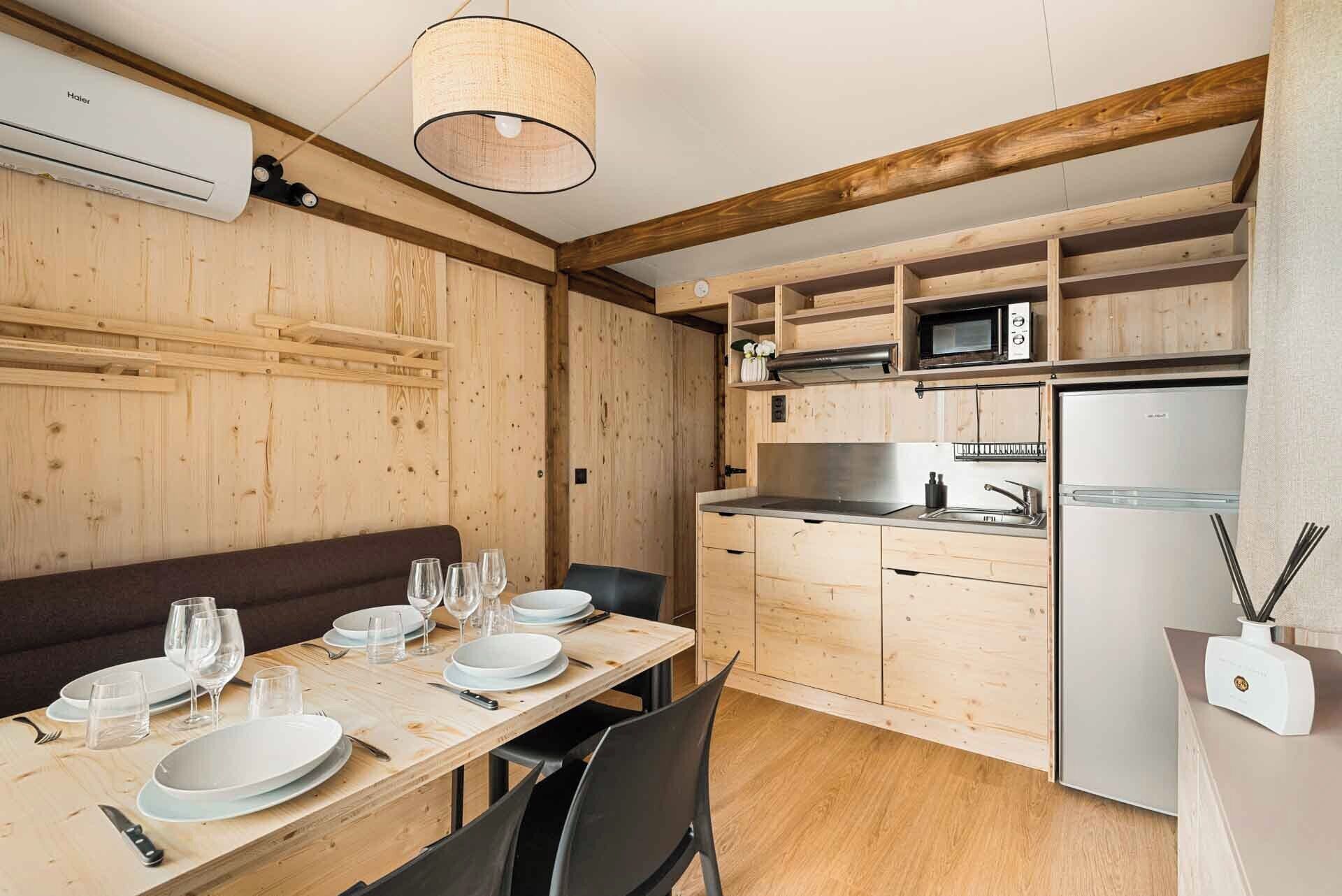 Interior em madeira com mesa de jantar posta para quatro, cozinha com armários de madeira, micro-ondas e geladeira.