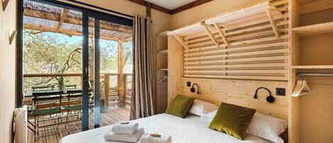 A bedroom with a large bed, a wooden headboard, a window with a view of trees, and a balcony with a table and chairs.