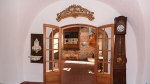 Puerta de madera abierta que da a un comedor con pared de piedra y una escalera de madera. Se ve un reloj de pie a la derecha.