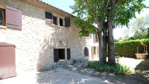 Un edificio de piedra con patio de grava, contraventanas de madera y un gran árbol.