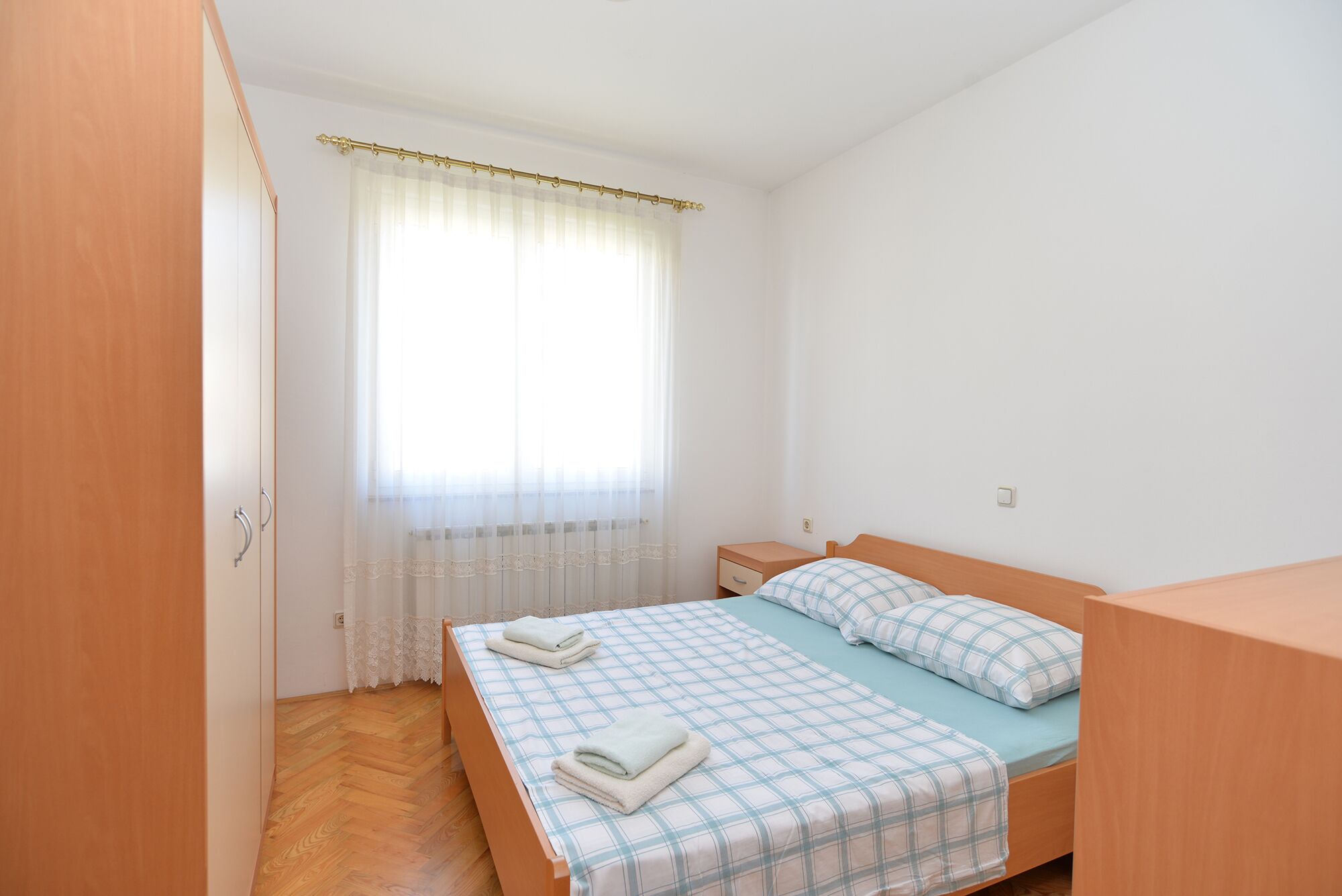 A bedroom with a wooden bed, a nightstand, a window with sheer curtains, and a wooden wardrobe.