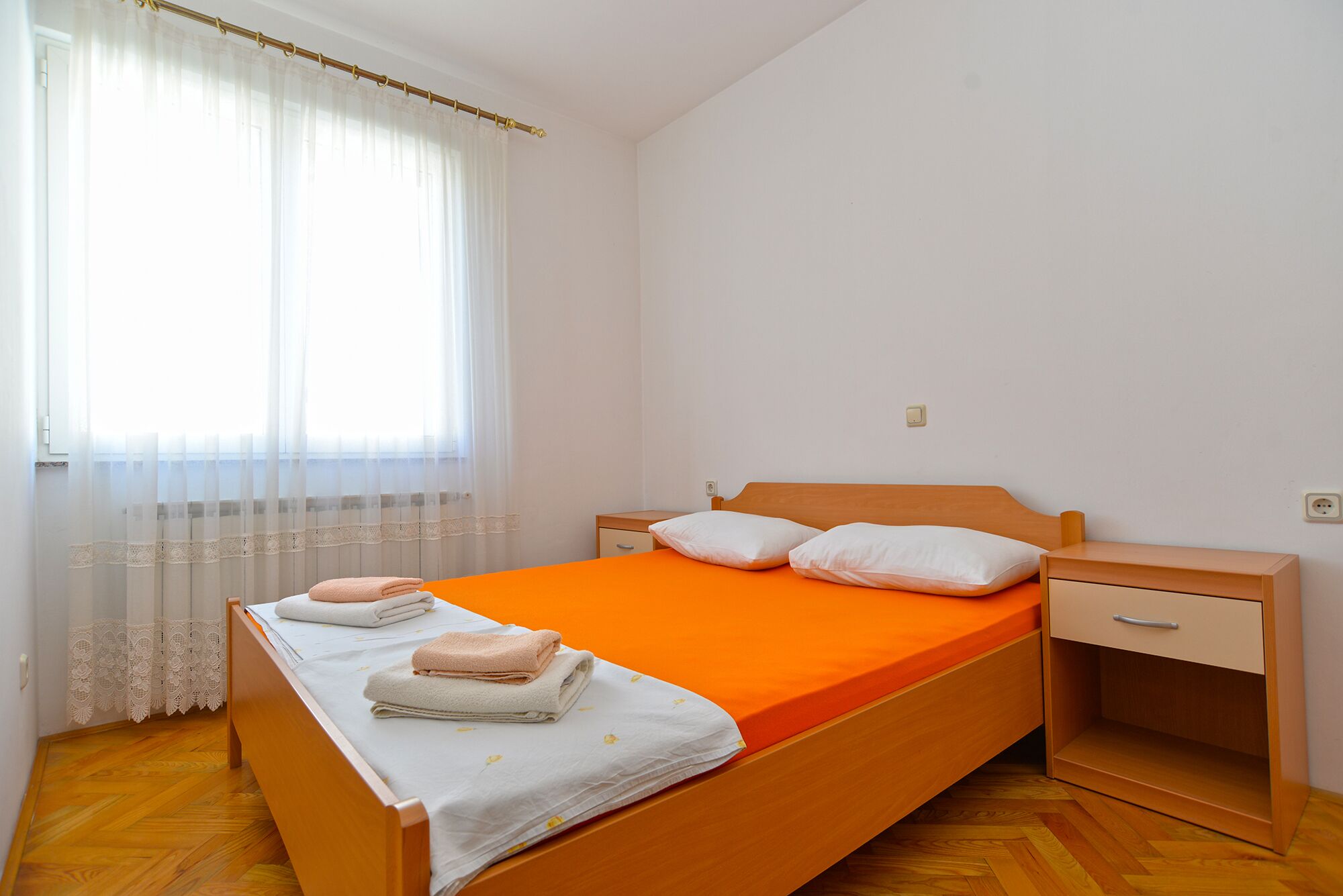 A single-bed room with a wooden headboard, white and orange bedding, a nightstand, and a small bedside table.