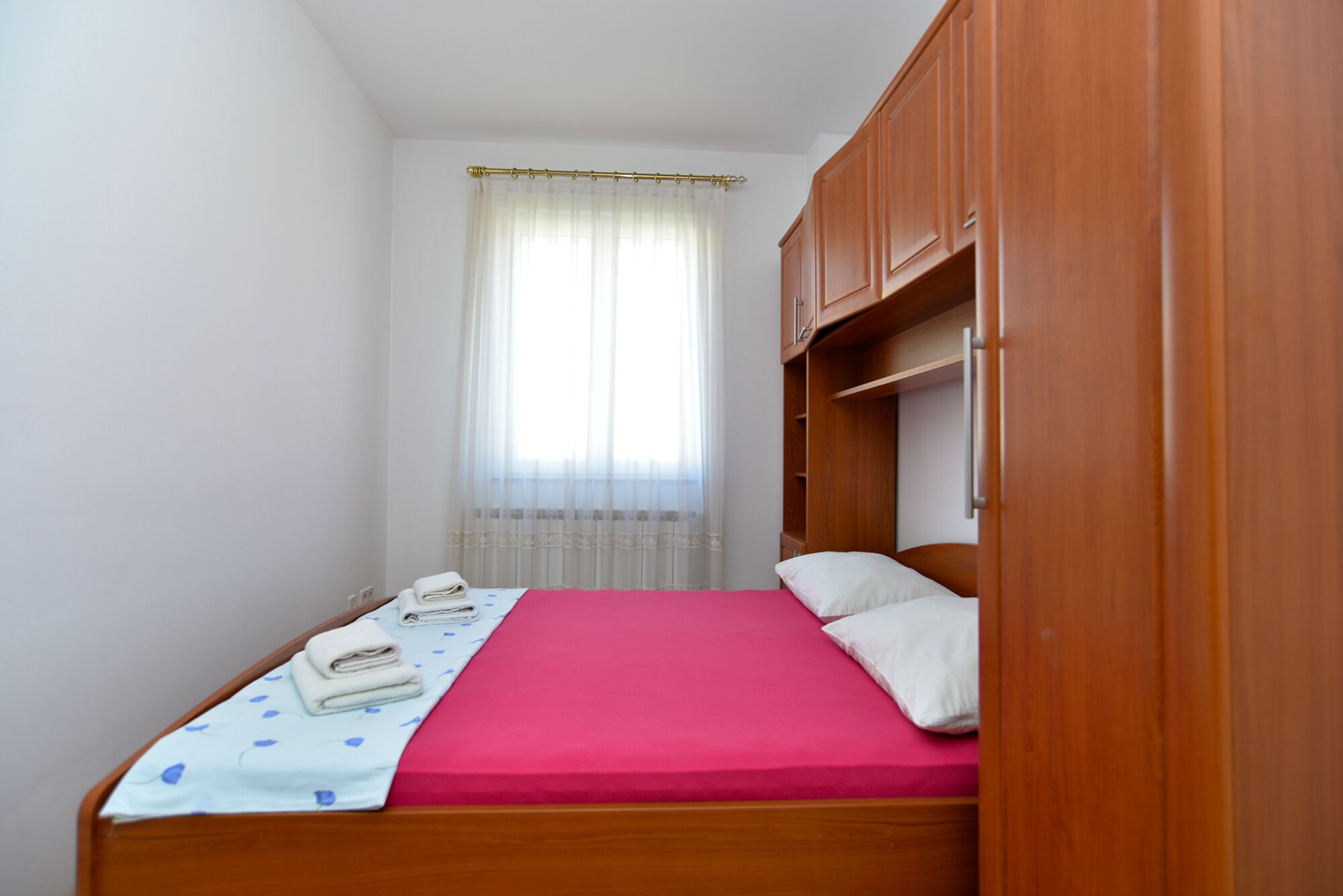 A single bed with a red bedspread, white pillows, and a folded towel on a white bedspread with blue patterns. There is a wooden wardrobe with a mirror and a window with sheer curtains.