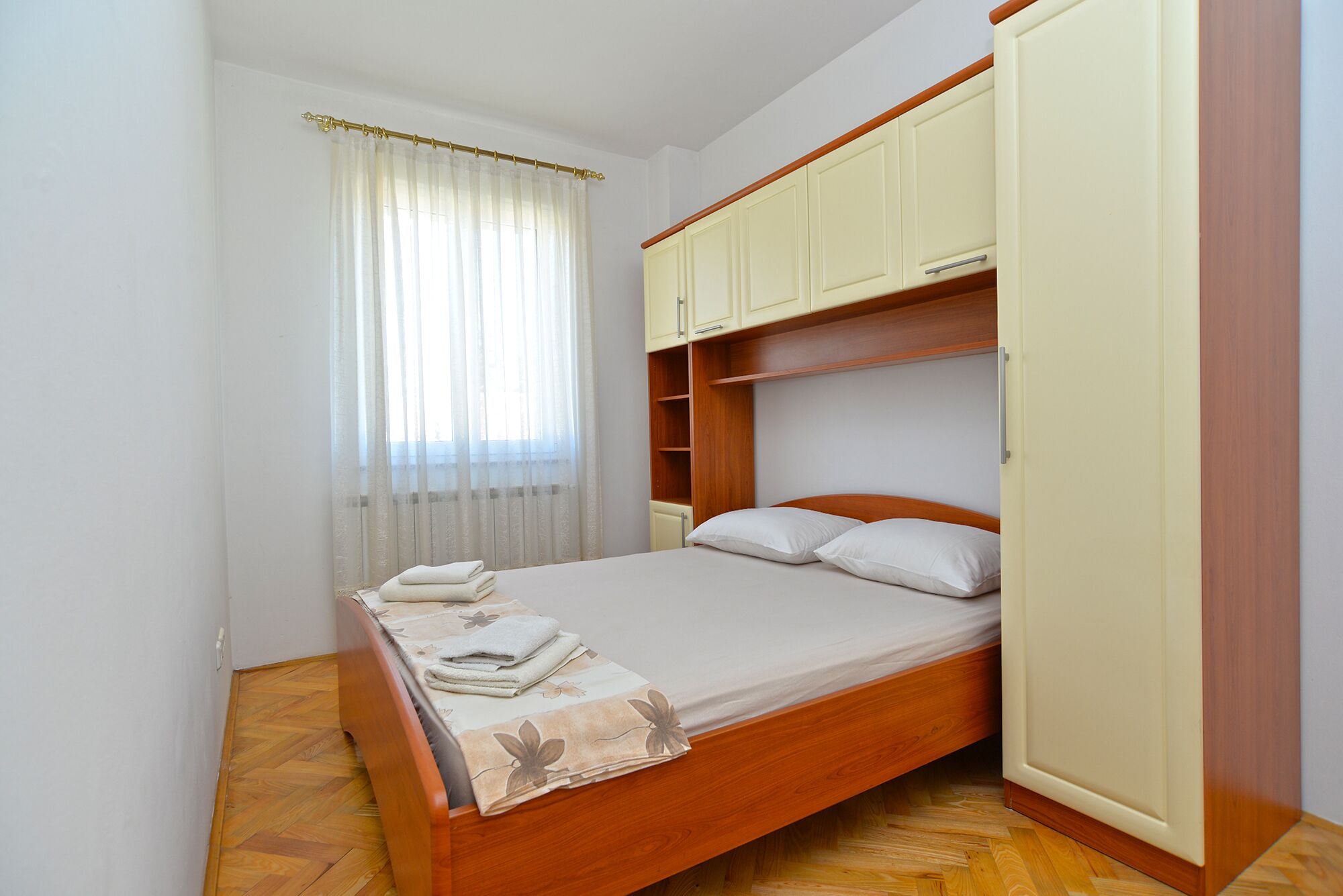 A compact bedroom with a wooden bed, a built-in wardrobe, and a window with sheer curtains.