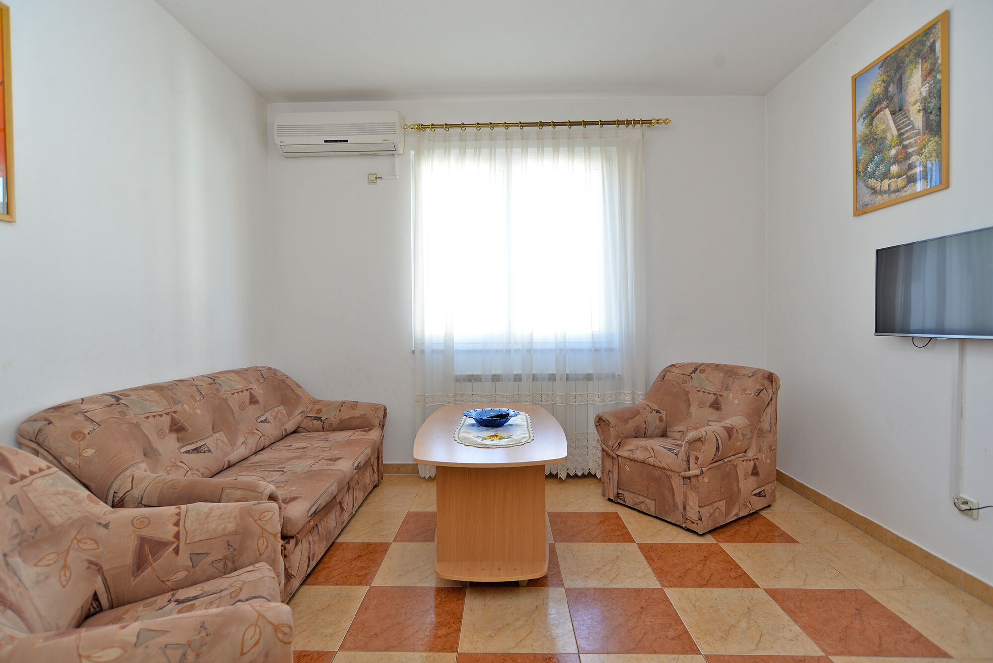 Una sala de estar con un sofá, un sillón y una mesita baja con un cuenco. Se ve una ventana con cortinas traslúcidas y un equipo de aire acondicionado en la pared.