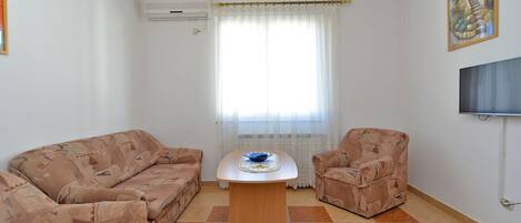 Una sala de estar con un sofá, un sillón y una mesita baja con un cuenco. Se ve una ventana con cortinas traslúcidas y un equipo de aire acondicionado en la pared.
