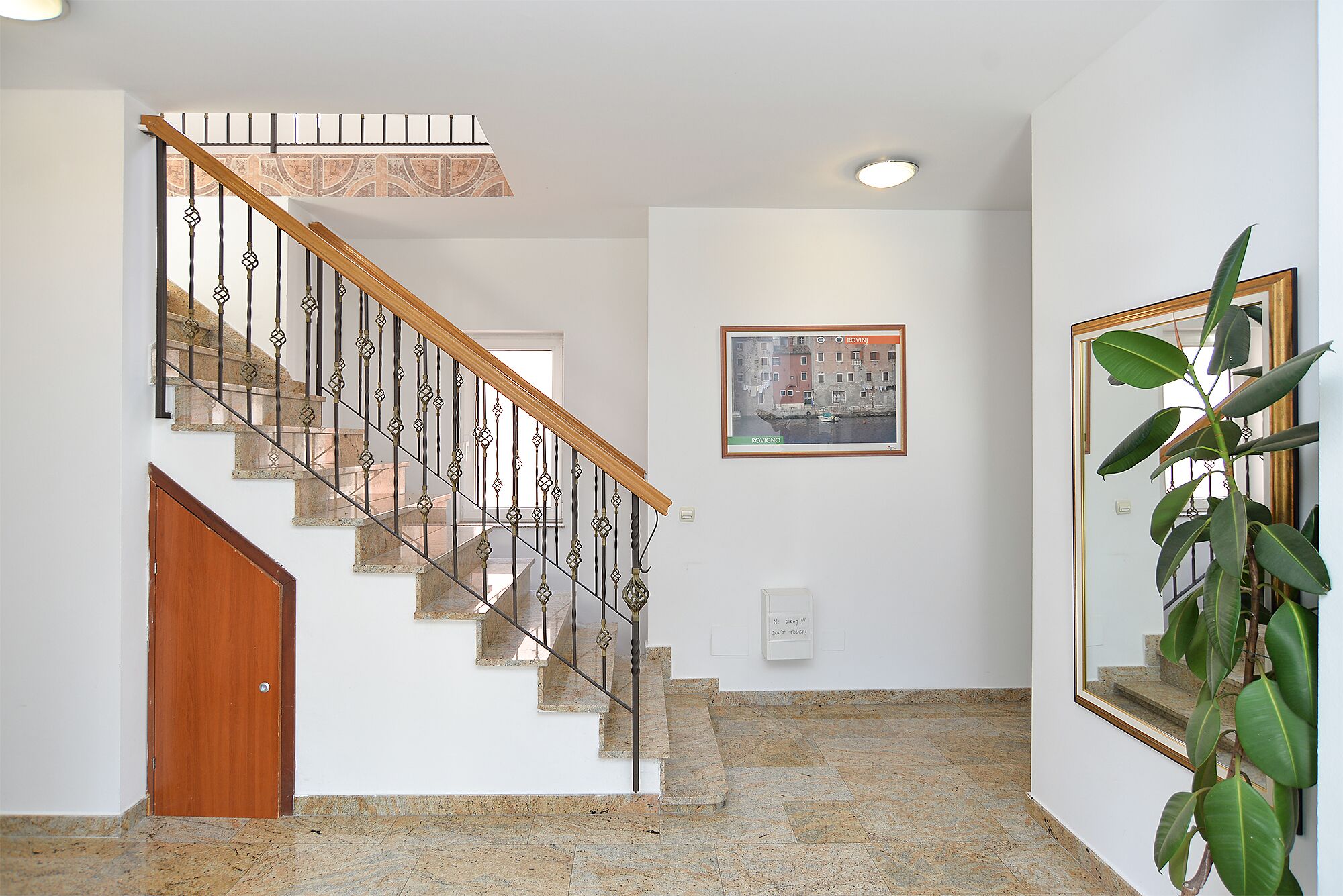 A staircase with a wooden handrail and metal balusters, leading to a landing with a framed picture and a potted plant.