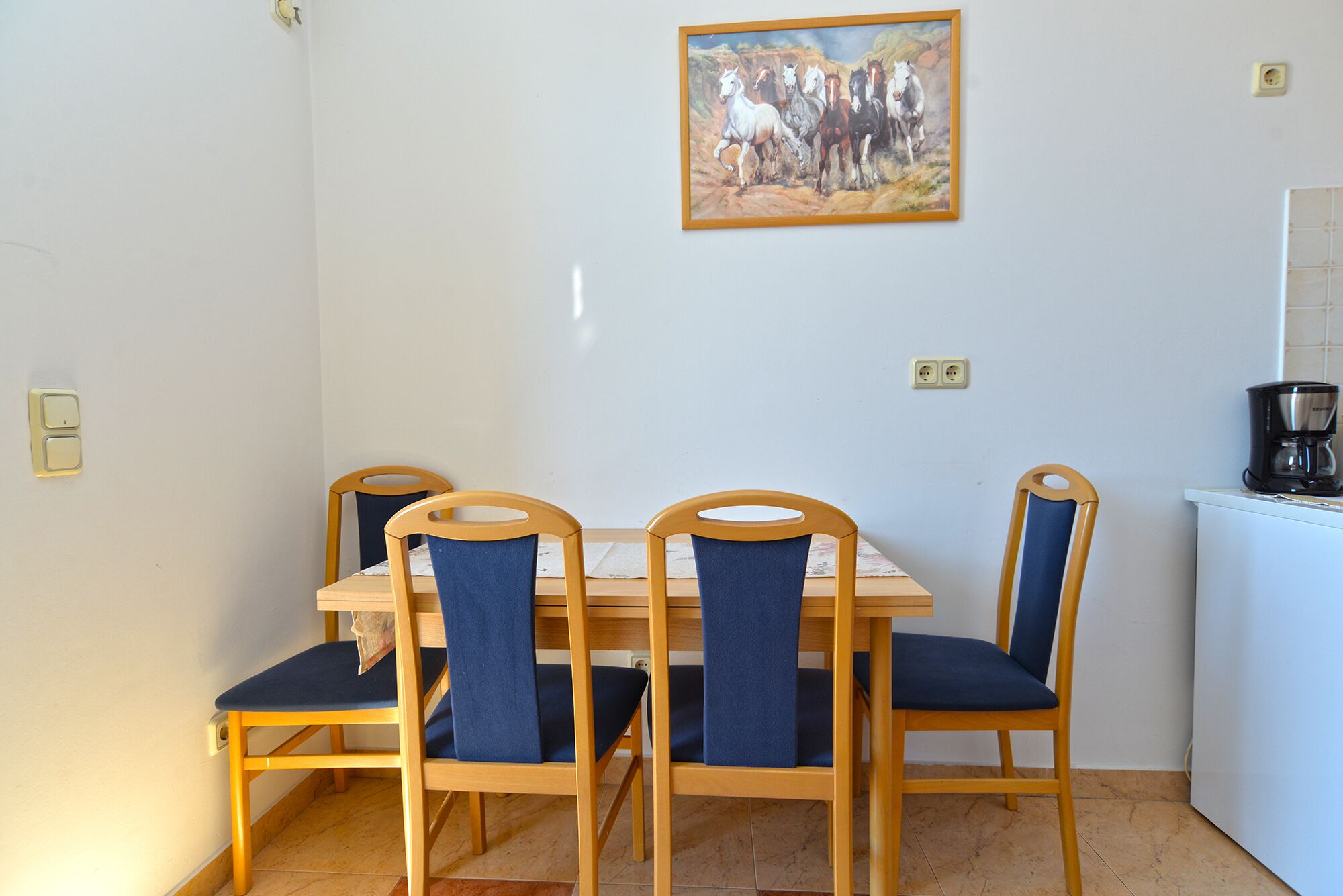 Ein Essbereich mit einem Holztisch, vier Stühlen, einem Bild an der Wand und einer Kaffeemaschine auf der Arbeitsfläche.