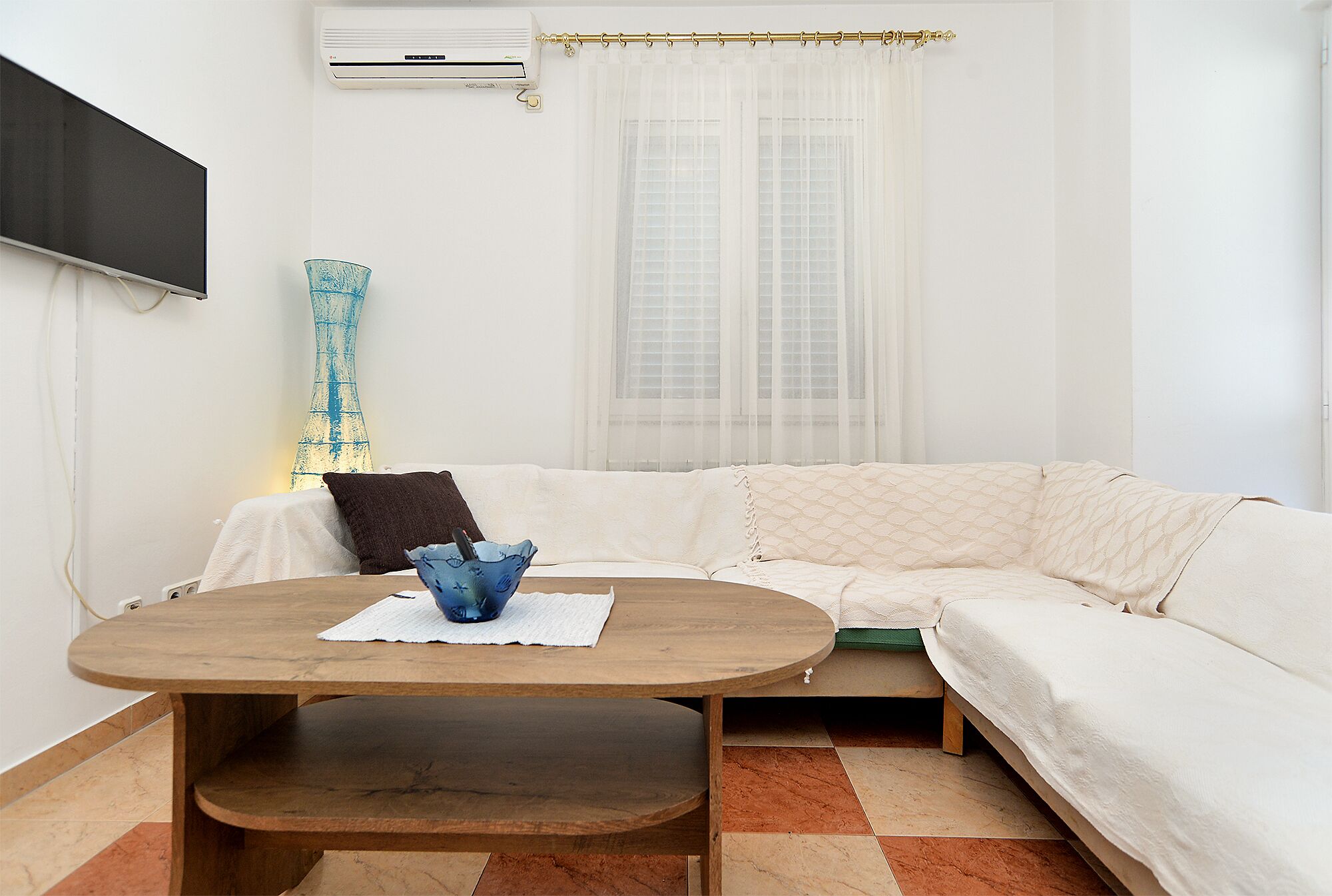 A modern living room with a white sofa, a wooden coffee table, a blue vase, and a television mounted on the wall.