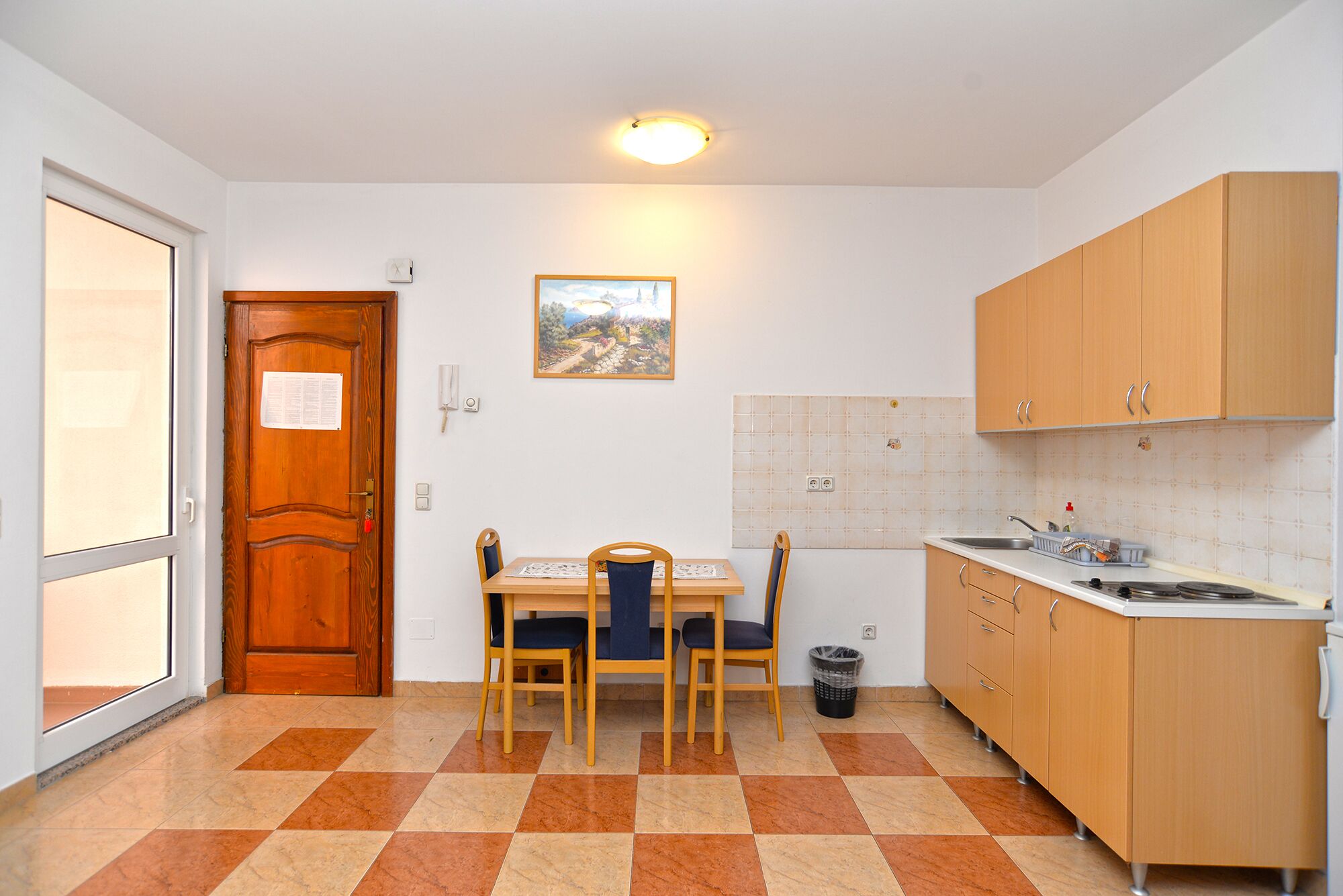 Una cocina compacta con armarios de madera, protector contra salpicaduras de azulejos, una mesa de comedor con sillas y un cesto de basura.