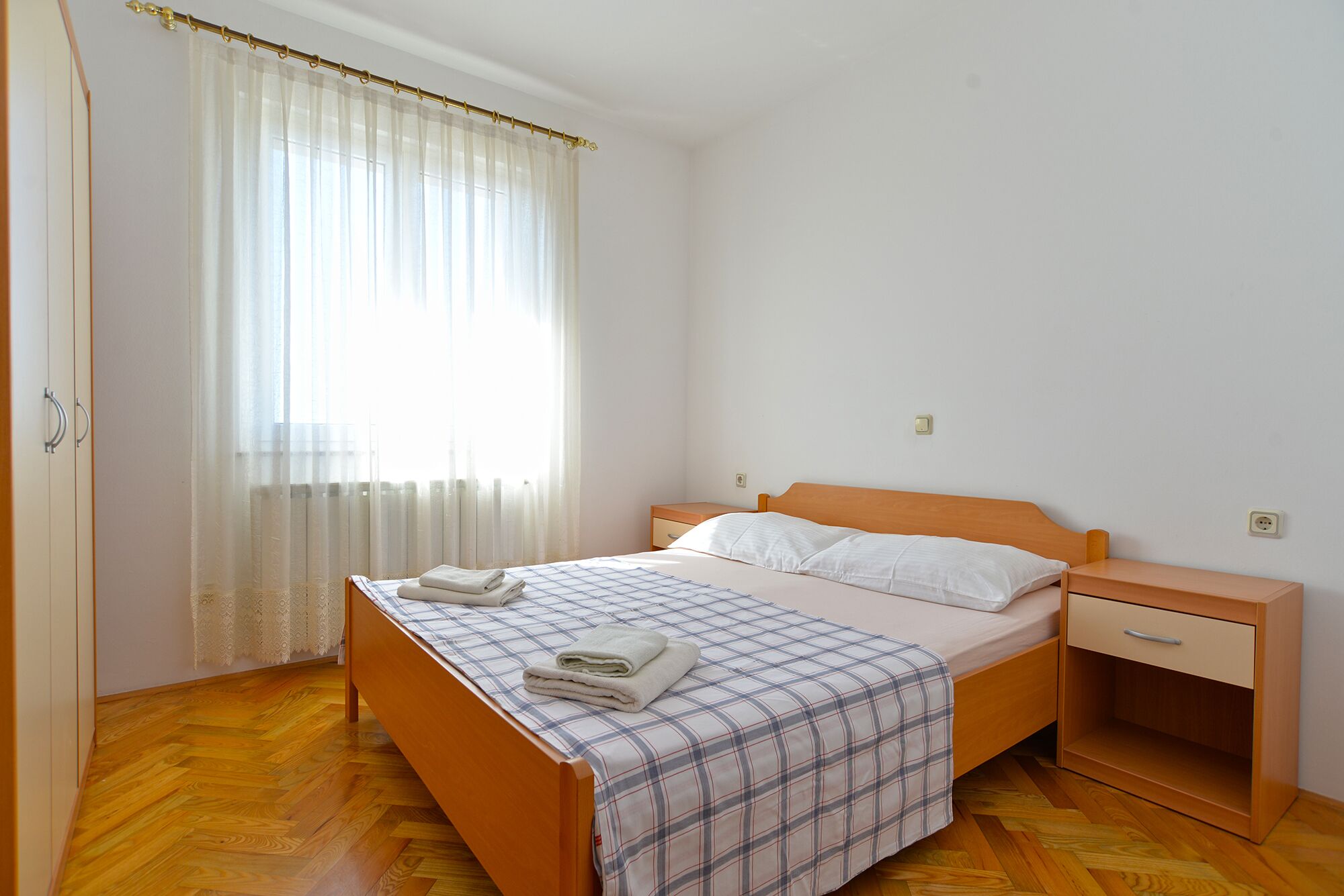 A bedroom with a wooden bed, a nightstand, a bedside table, and a window with sheer curtains.