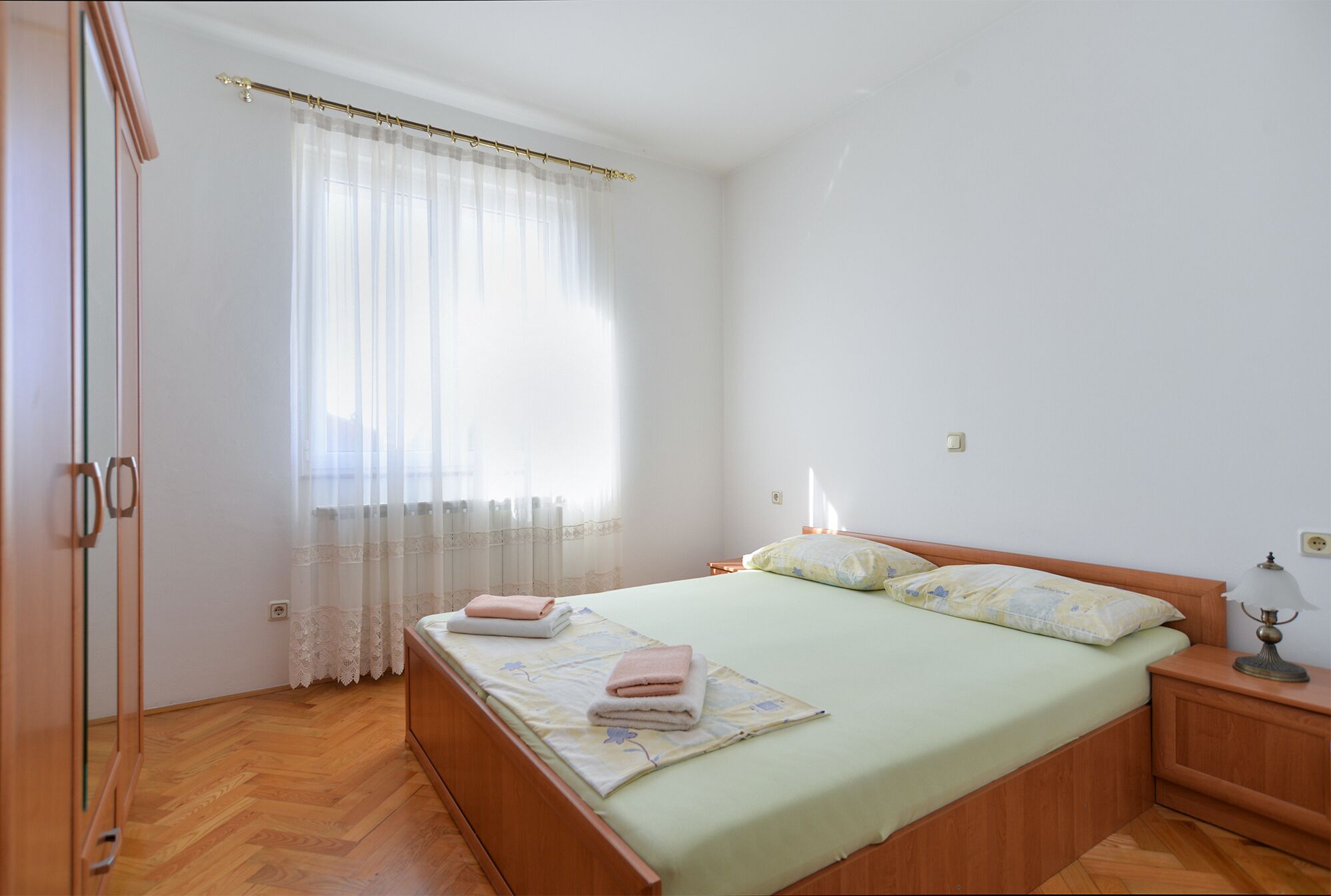A bedroom with a bed, a nightstand, a lamp, a window with sheer curtains, and a wooden wardrobe.