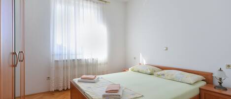 A bedroom with a bed, a nightstand, a lamp, a window with sheer curtains, and a wooden wardrobe.