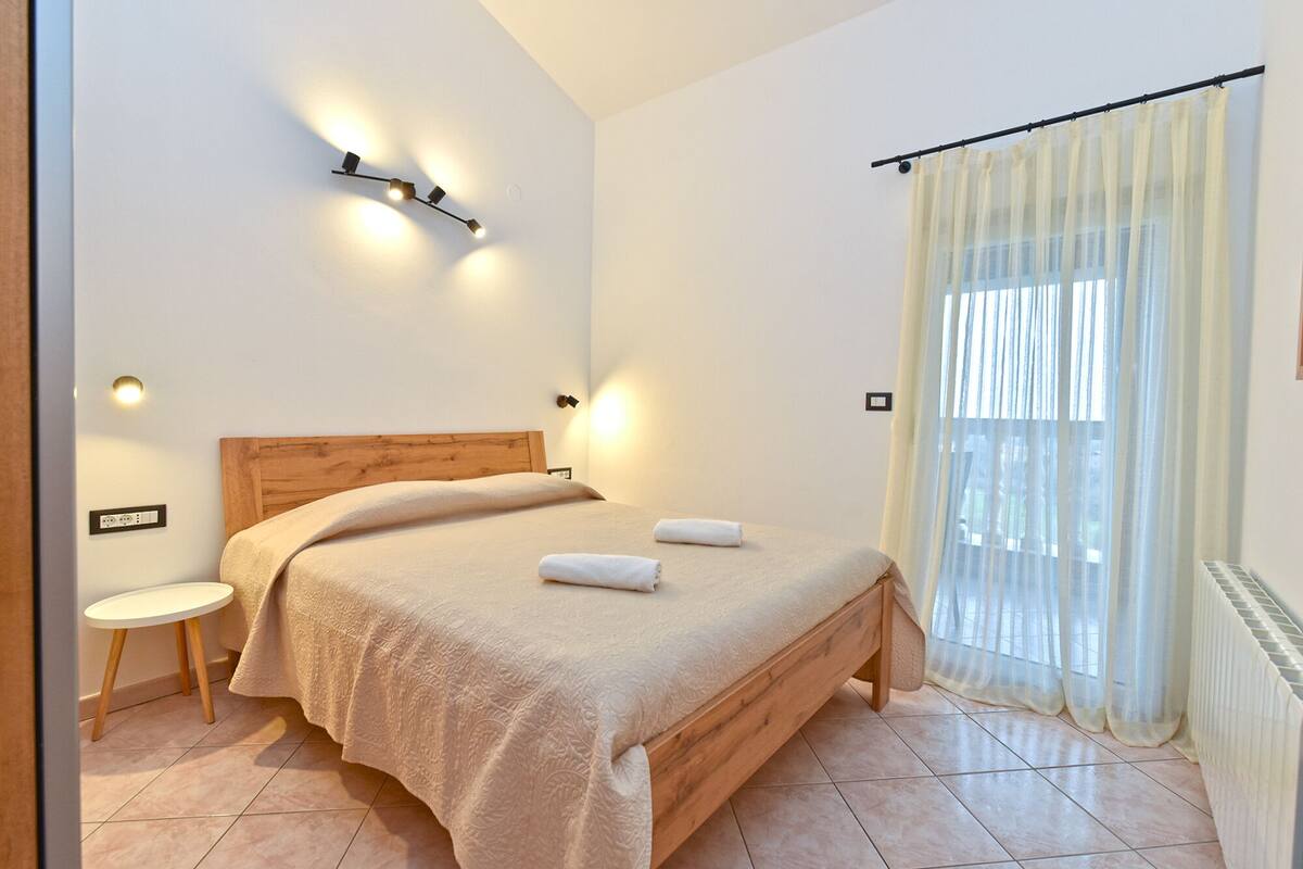 A bedroom with a wooden bed, a bedside table, a wall-mounted light fixture, and a window with sheer curtains.