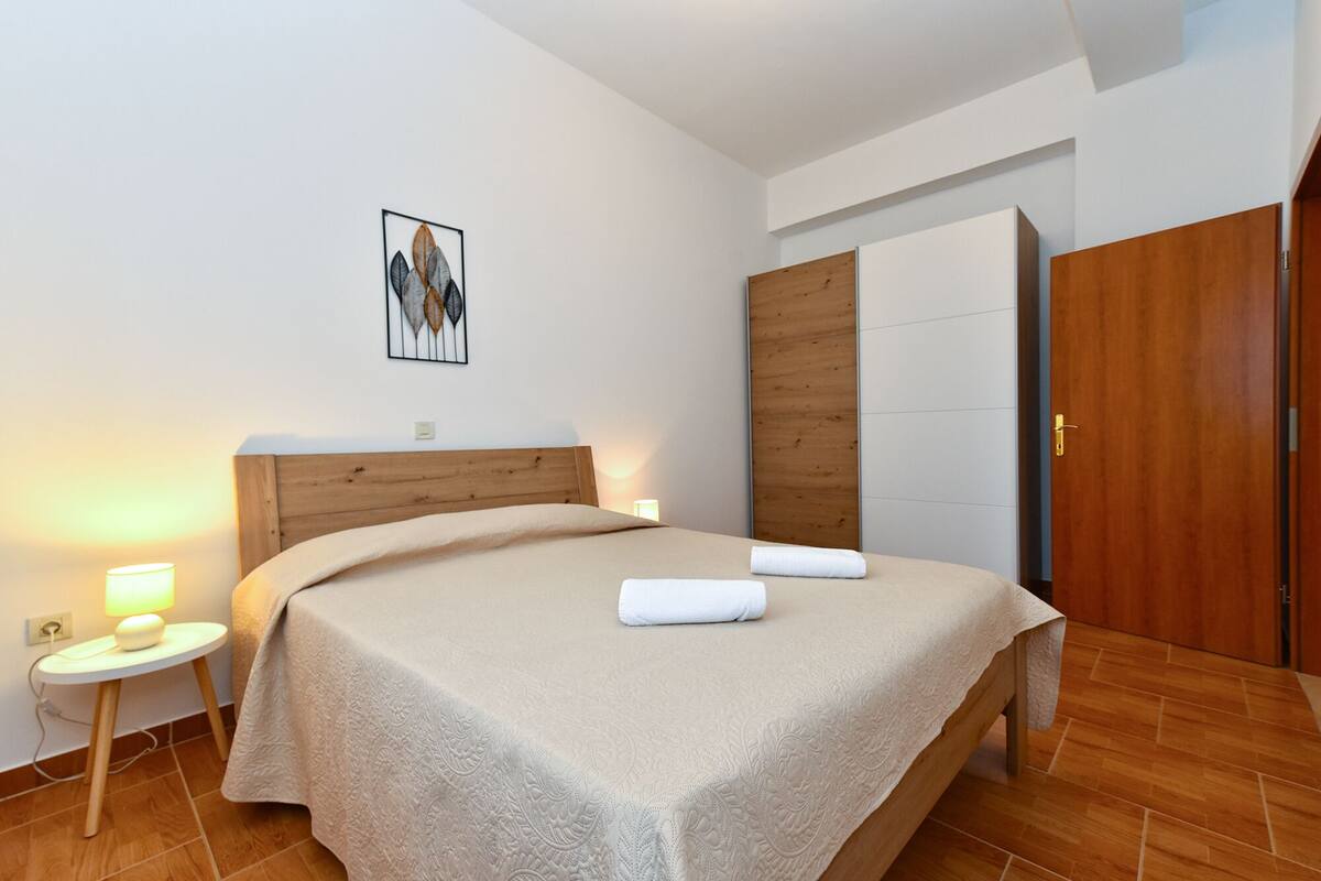 A hotel room with a wooden headboard, a large bed with a textured white bedspread, a bedside table with a lamp, and a wooden wardrobe.