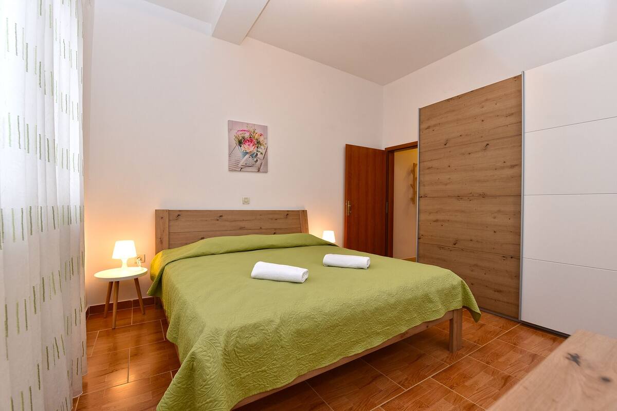 A bedroom with a wooden headboard, a green bedspread, two white towels, a bedside lamp, and a wooden wardrobe.
