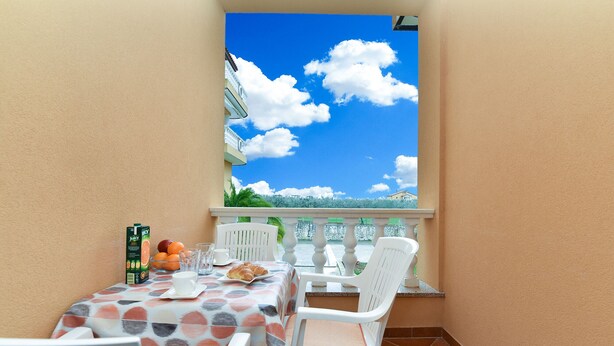 Un balcón con una mesa para dos, mantel con estampado de lunares, sillas blancas y vista a un edificio y al cielo a través de una ventana.