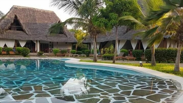 A swimming pool with a mosaic tile design, surrounded by palm trees and a thatched-roof building. - JOHARI VILLA (Malindi)