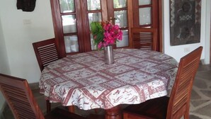 A round wooden dining table with a patterned tablecloth, surrounded by wooden chairs. A hanging light fixture is above the table, and a vase with flowers is placed on it. - JOHARI VILLA (Malindi)
