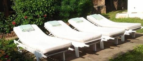 A row of white sun loungers with towels, set against a backdrop of lush greenery and a thatched-roof structure.