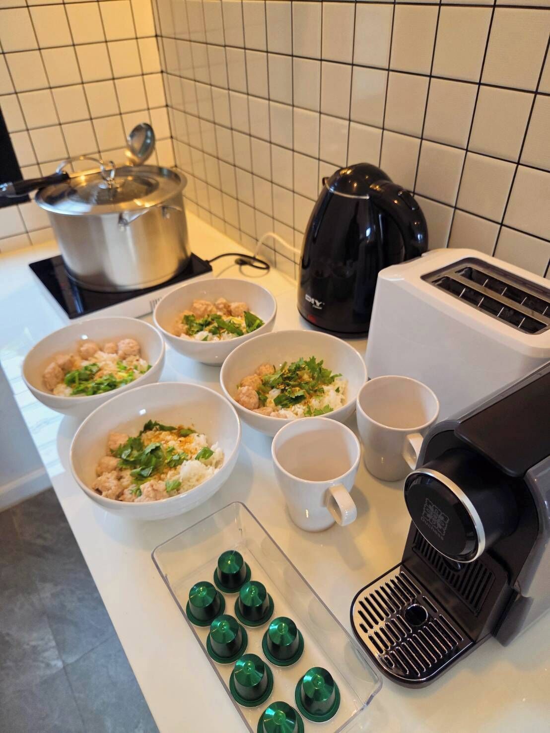 Egy konyha fehér csempékkel, tűzhellyel, vízforralóval, pirítóval és tálakban tálalt ételekkel.