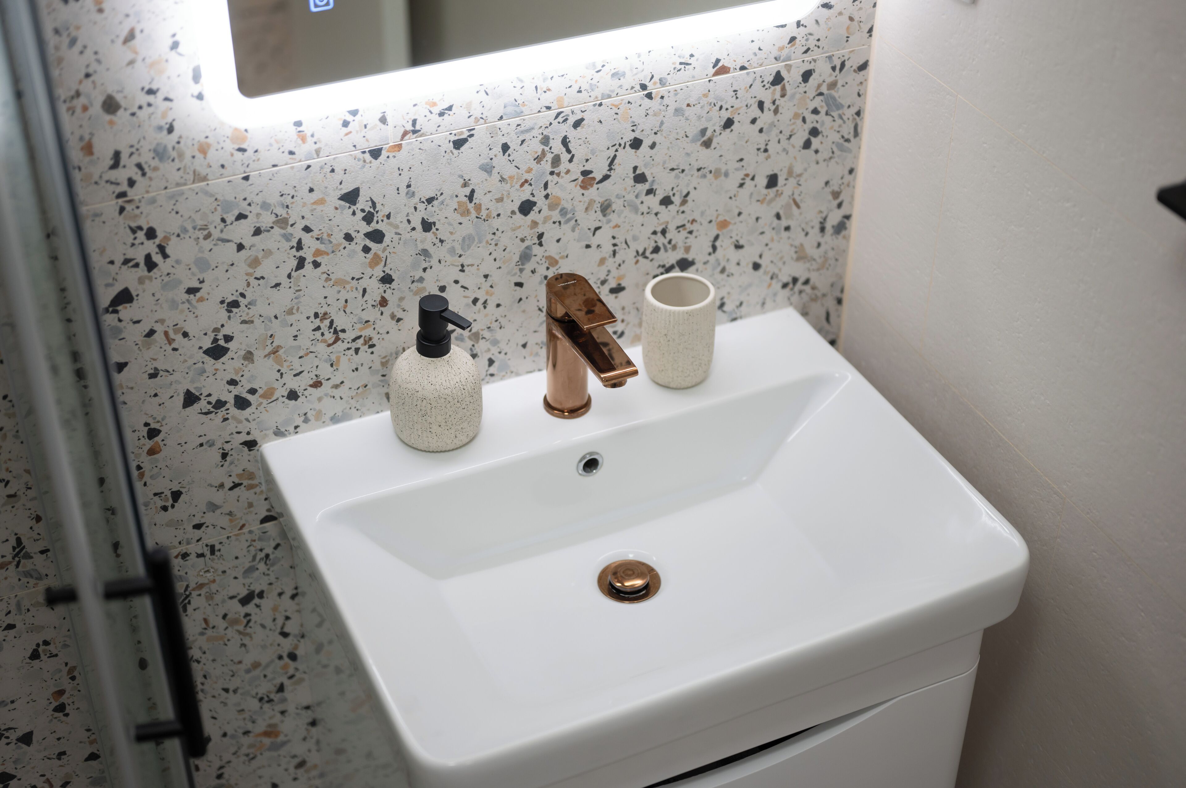 A bathroom with a white sink, a copper faucet, a soap dispenser, and a cup.