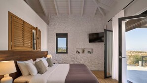 Ein Schlafzimmer mit Steinwand, einem Holzkopfteil, einem Bett mit weißer Bettwäsche, einer Nachttischlampe, einem an der Wand montierten Fernseher und einer Glastür zum Außenbereich.