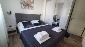 Quarto moderno com uma cama grande, cabeceira estofada cinza, mesa de cabeceira, luminária de parede e um espelho amplo.