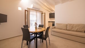 A modern living room with a sofa, a dining table with chairs, a TV mounted on the wall, and a kitchen area in the background.