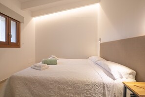 Een keurig opgemaakt bed met een witte deken en kussens, een houten nachtkastje en een raam met zonwering.